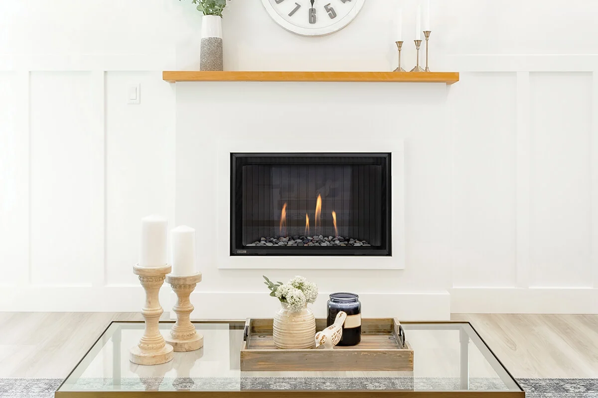 Montigo DelRay Square gas fireplace set into a white board-and-batten wall with a light oak mantel, candlesticks and a wall clock above, in a bright living room