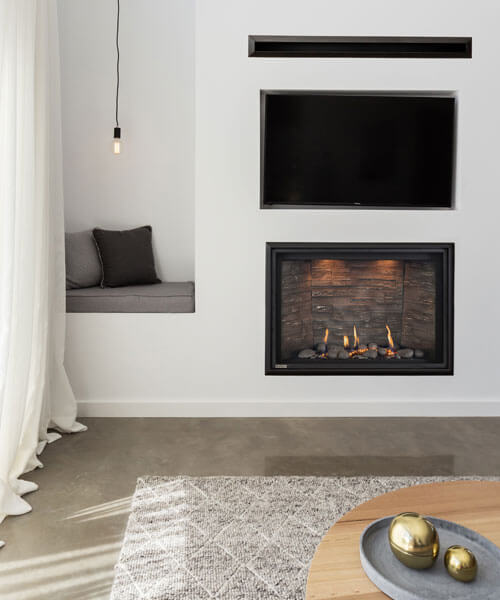 Montigo DelRay Square gas fireplace with a wood-look interior and stone media, set into a white wall below a recessed television beside a built-in window bench