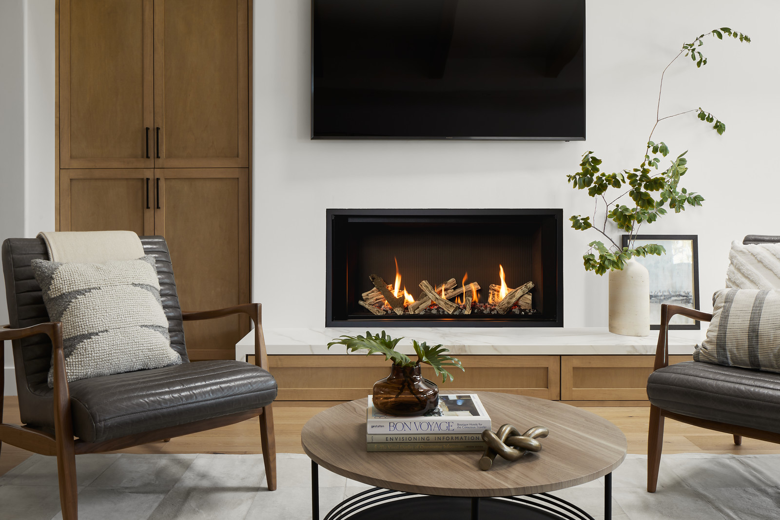 A tall, wide linear gas fireplace with a realistic split-log firebed and a television mounted on the wall above it in a bright modern living room