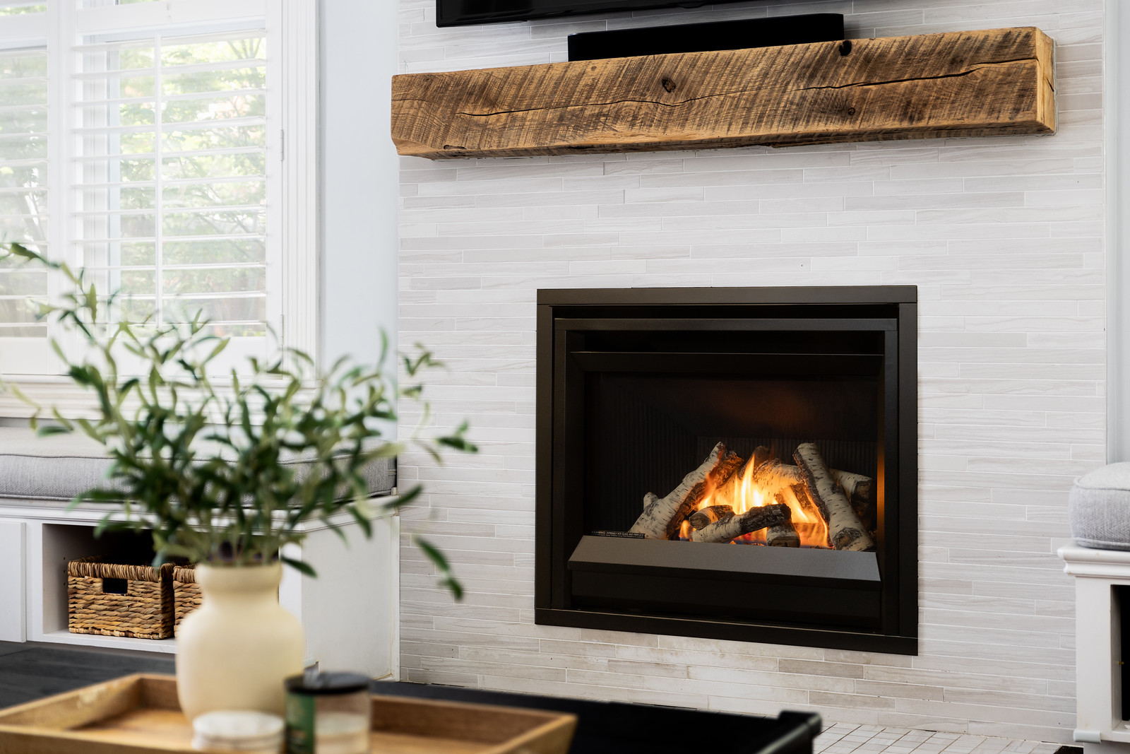 Valor H4 radiant gas fireplace with a birch log set and a black four-sided front, built into a white tile wall beneath a reclaimed-wood mantel