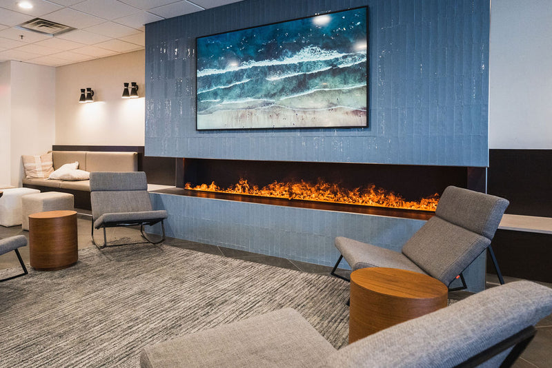 A long Aquafire water-vapour fireplace built into a tiled feature wall in a hotel lounge, a wide ribbon of flame beneath framed ocean art with lounge chairs alongside — an install where a real fire could never go
