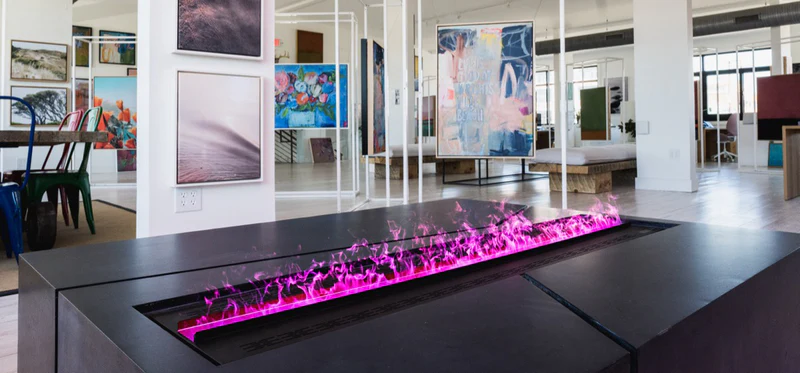 Aquafire linear fireplace in a black island console showing its LED colour range, the water-vapour flame glowing bright pink-magenta in a loft art gallery