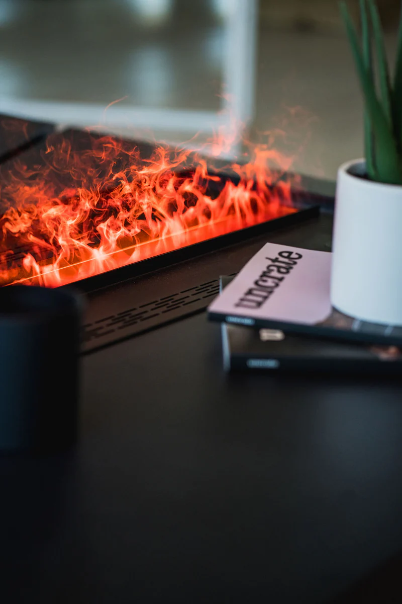 Close view of Aquafire's water-vapour flame — soft orange mist lit from below to look like curling fire — rising from a slim black linear unit on a console table beside a potted plant