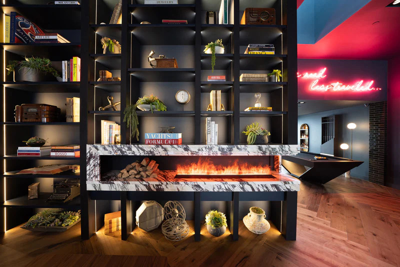 Aquafire linear water-vapour fireplace built into a marble shelf within a black bookshelf room divider, a wide band of flame-like lit mist over faux logs, in a design-forward lounge with a neon sign