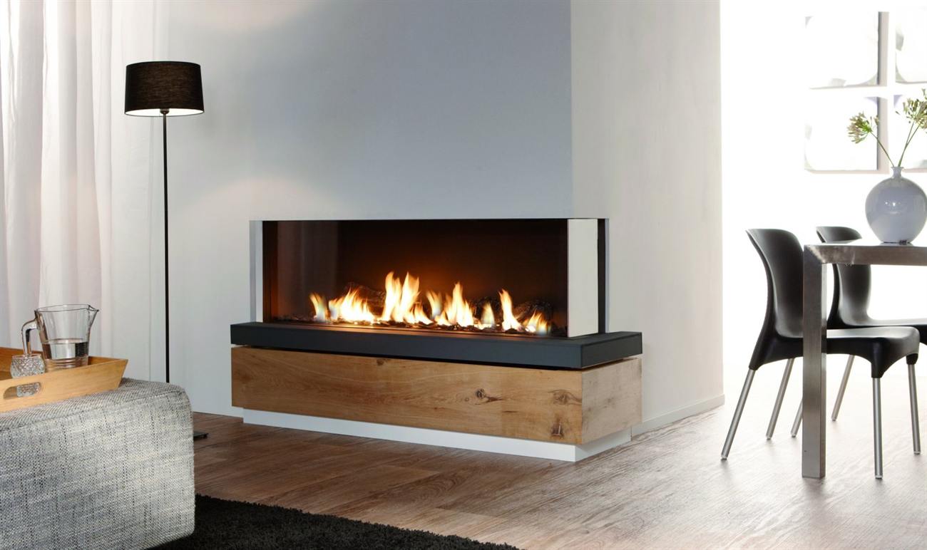 Element4 Bidore 140 gas fireplace built into a pale grey wall above a wide oak plinth, its glass wrapping the front and one return so the long low bed of flame is visible from the living room and the dining table beyond