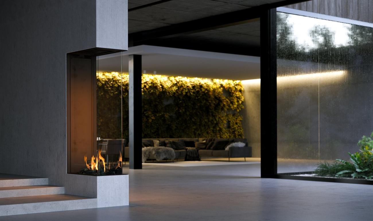 Element4 Sky Large Roomdivider gas fireplace built into a board-formed concrete pier as a tall portrait column of glass, flames rising from a dark stone bed, with a covered terrace and a lit green wall visible through the fire and rain running down the glazing beyond