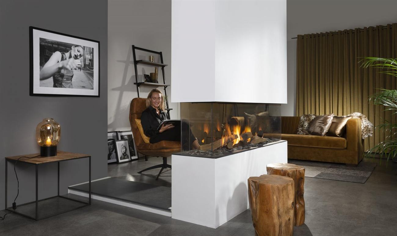 Element4 Summum 140 4/S four-sided gas fireplace standing as a free-standing column in the middle of a living room, glazed on every face above a white plinth, with a tan leather armchair on one side and a velvet sofa on the other, both facing the same fire