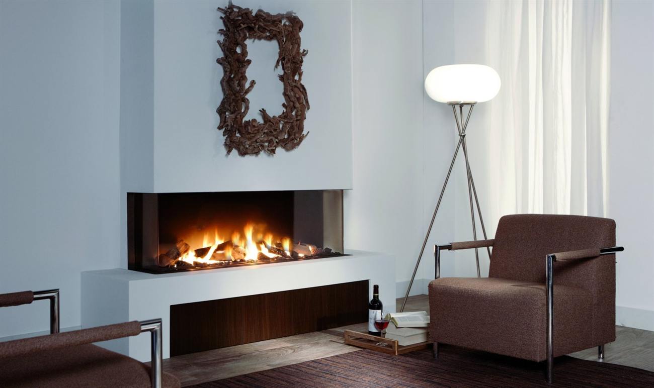 Element4 Trisore 140 three-sided gas fireplace projecting from a white plaster chimney breast above a dark timber plinth, glass on the front and both ends so the flames over the log bed are visible from across the room and from either side