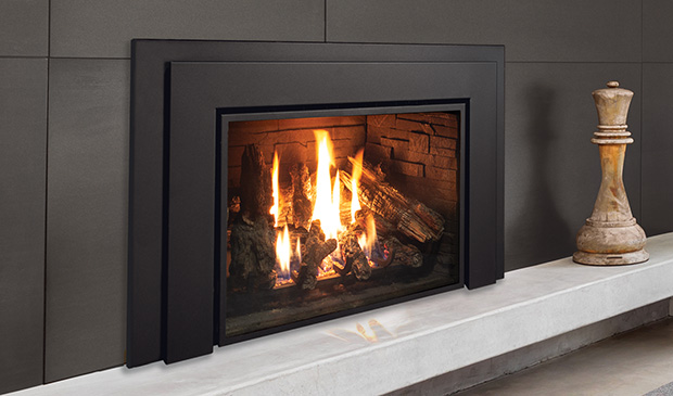 Enviro E44 gas insert with a broad black contemporary surround set into a charcoal wall above a white stone bench hearth, a large log fire behind the glass, a giant carved chess piece beside it