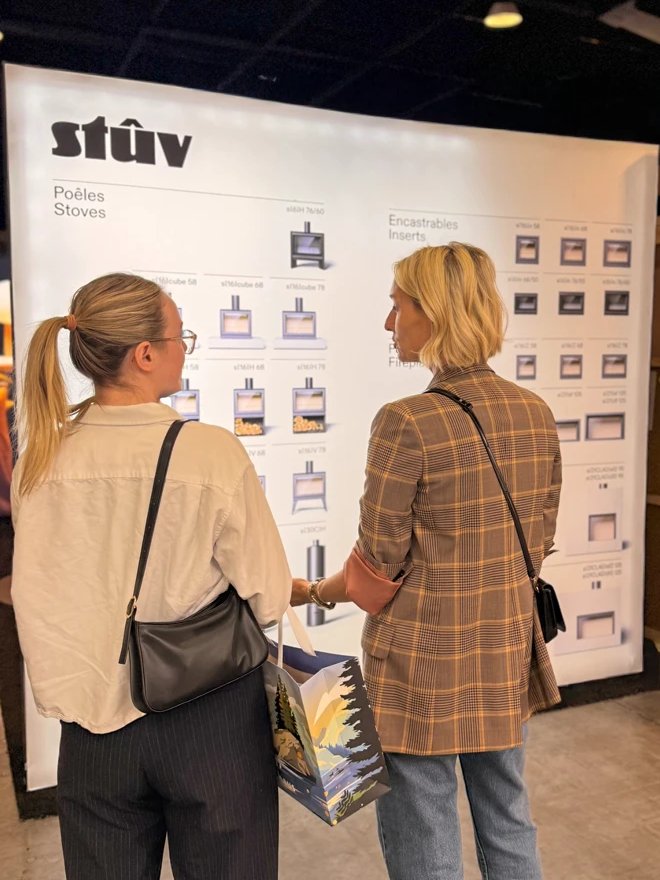 Two women study the Stûv display wall of stoves and inserts at IDS Vancouver 2026; on the right, in a plaid blazer, is Meredith Cormack, Vice President of Maxwell Fireplace