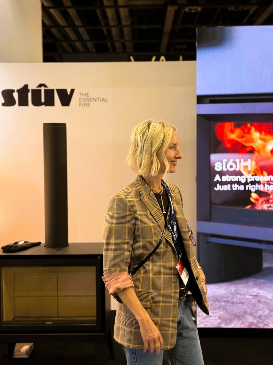 Meredith Cormack, Vice President of Maxwell Fireplace, smiling at the Stûv stand in front of a matte-black Stûv stove and a screen showing the Stûv s[6]H
