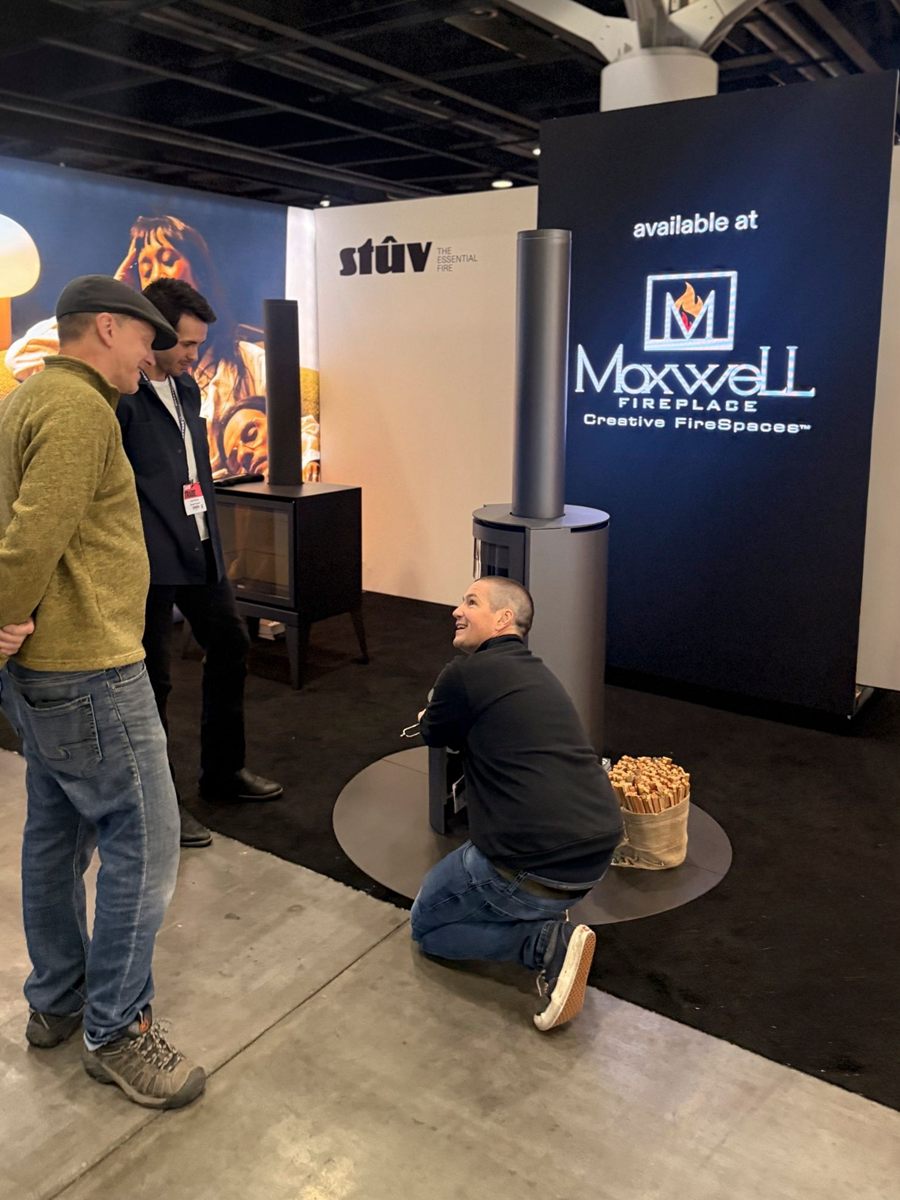 Samuel Choinière-Deslauriers of Stûv in conversation beside a cylindrical Stûv stove at the IDS Vancouver 2026 stand, beneath an illuminated sign reading “available at Maxwell Fireplace, Creative FireSpaces™”