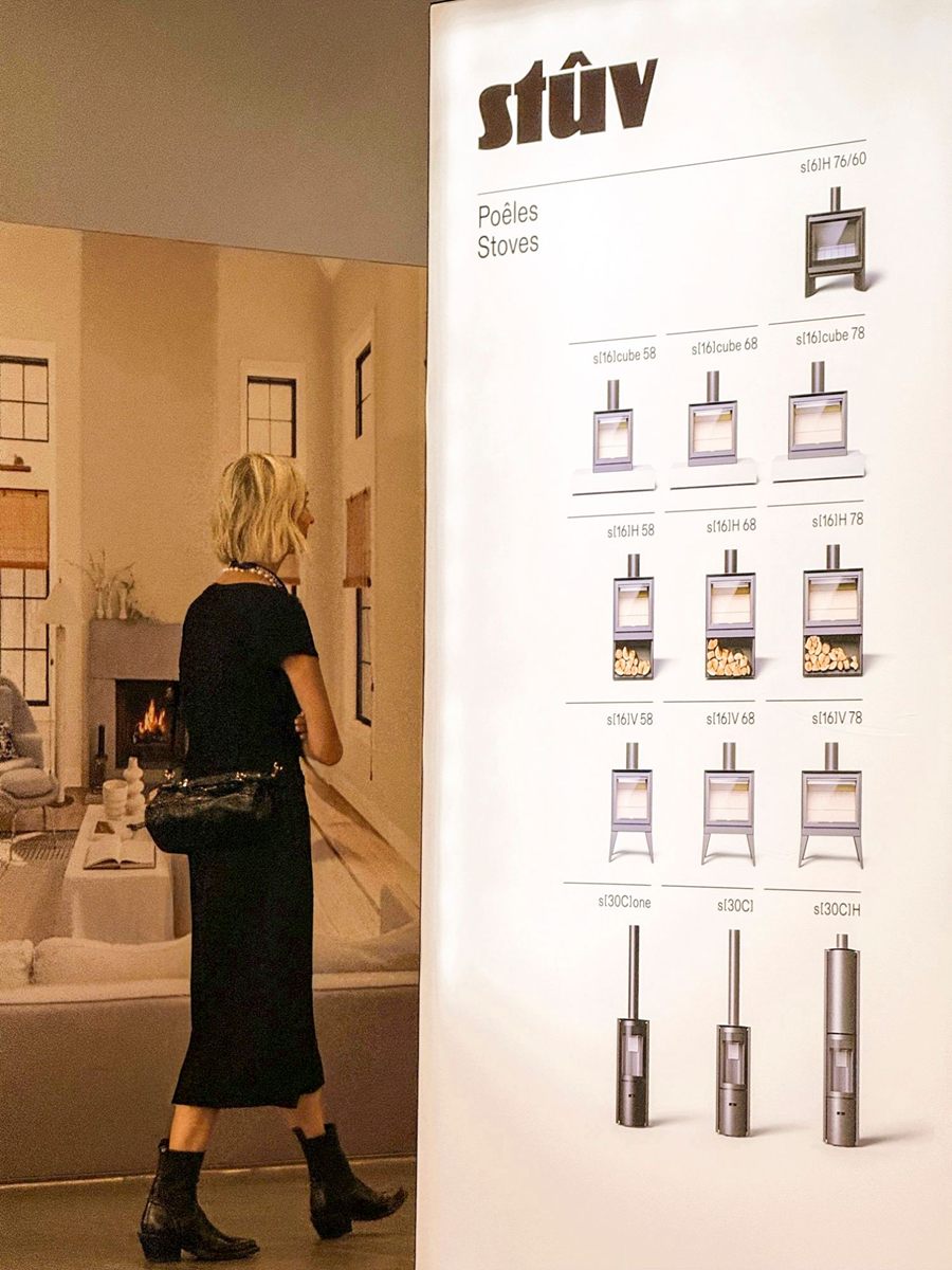A woman in a black dress walks past the Stûv stove lineup board at IDS Vancouver 2026, beside a large print of a sunlit living room with a fire burning