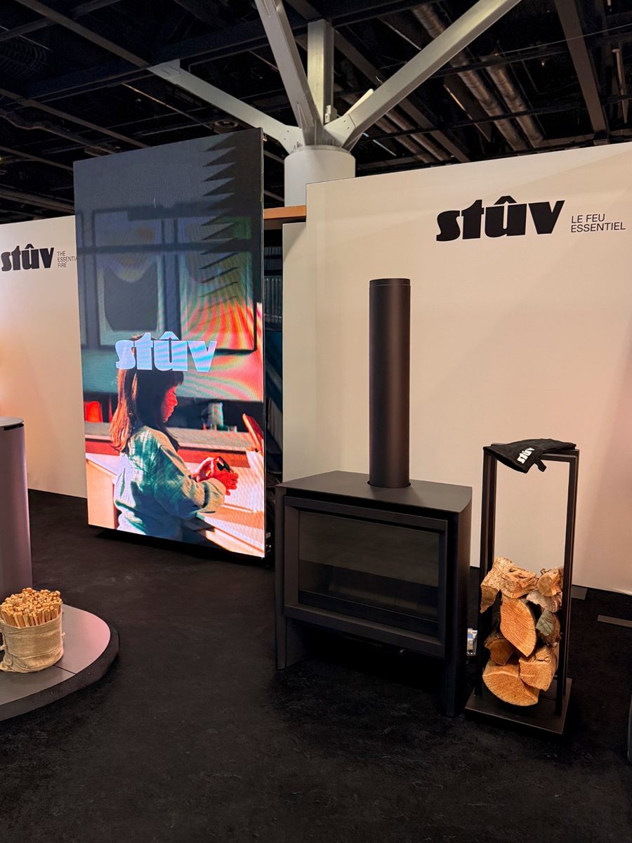 The Stûv stand at IDS Vancouver 2026: a matte-black freestanding Stûv wood stove with a tall flue, a log holder beside it, and a large video screen under the Stûv logo