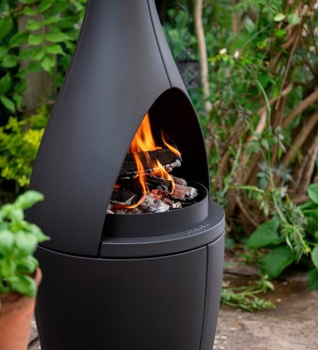 Close-up of the Morsø Kamino outdoor fireplace in a garden setting, with flames burning inside the arched cast iron opening and basil and herbs in pots alongside