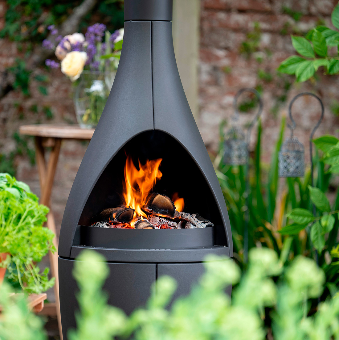 Morsø Kamino outdoor fireplace lit against a brick garden wall, with a bouquet of flowers in a vase and hanging lanterns alongside fresh greenery