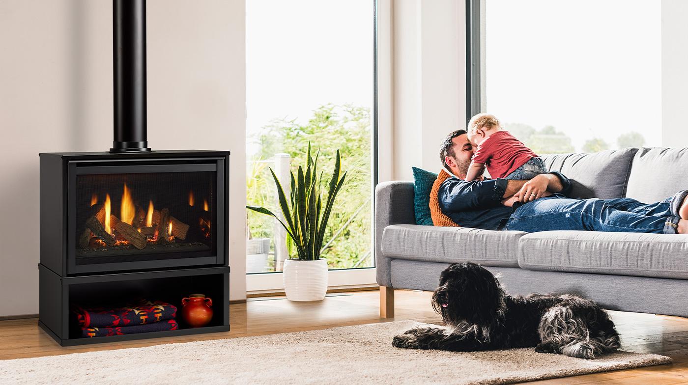 Majestic Ruby freestanding gas fireplace with a black front and a log fire beside a tall window, a man on a grey sofa lifting a toddler, and a black dog resting on the rug in front