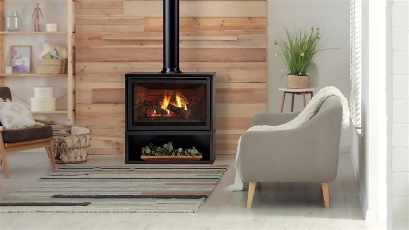 Majestic Trilliant freestanding gas fireplace on its black pedestal, a log fire behind the glass and a black flue rising to the ceiling, in a living room with a wood-slat wall and a grey armchair