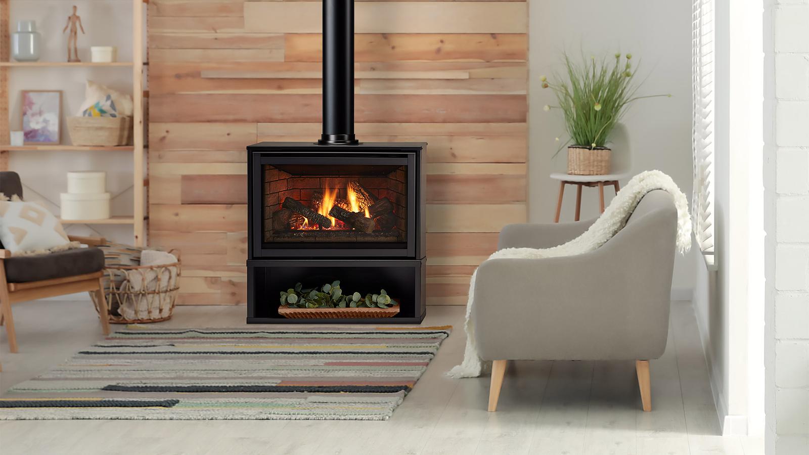 Majestic Trilliant freestanding gas fireplace on its black pedestal, a log fire in front of brick-pattern Canyon Brown panels and a black flue rising to the ceiling, in a living room with a wood-slat wall, a grey armchair and a striped rug