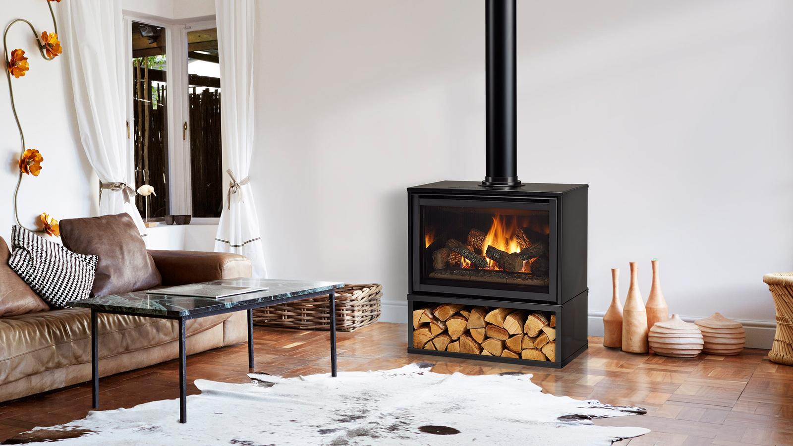 Majestic Trilliant freestanding gas fireplace beside a leather sofa on a parquet floor, its open lower shelf stacked with split logs and a black flue rising beside it