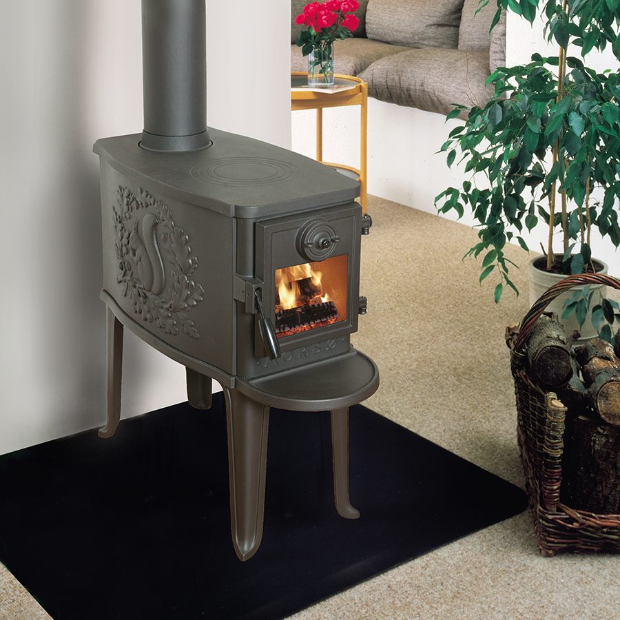 Morsø 2B Standard cast-iron wood stove on four tall legs with the cast relief on its side and a glass door, lit in a timber-clad room beside a sofa