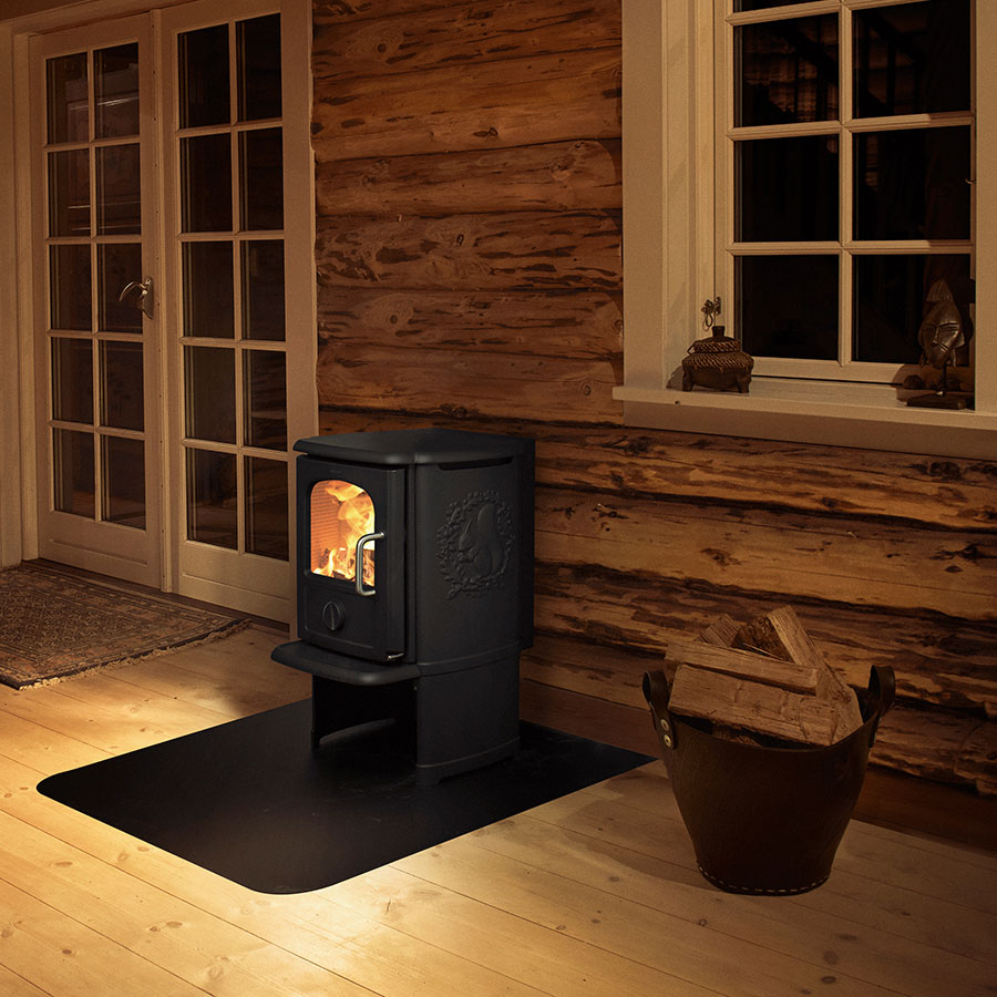 Morsø 3142 B cast-iron wood stove with the squirrel relief on its side, lit on a black floor plate in a timber-lined cabin room