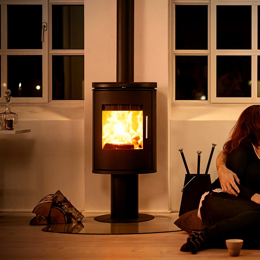Morsø 6148 B cast-iron wood stove standing on its slim pedestal, lit in a dark room beside a basket of logs and a set of fire tools