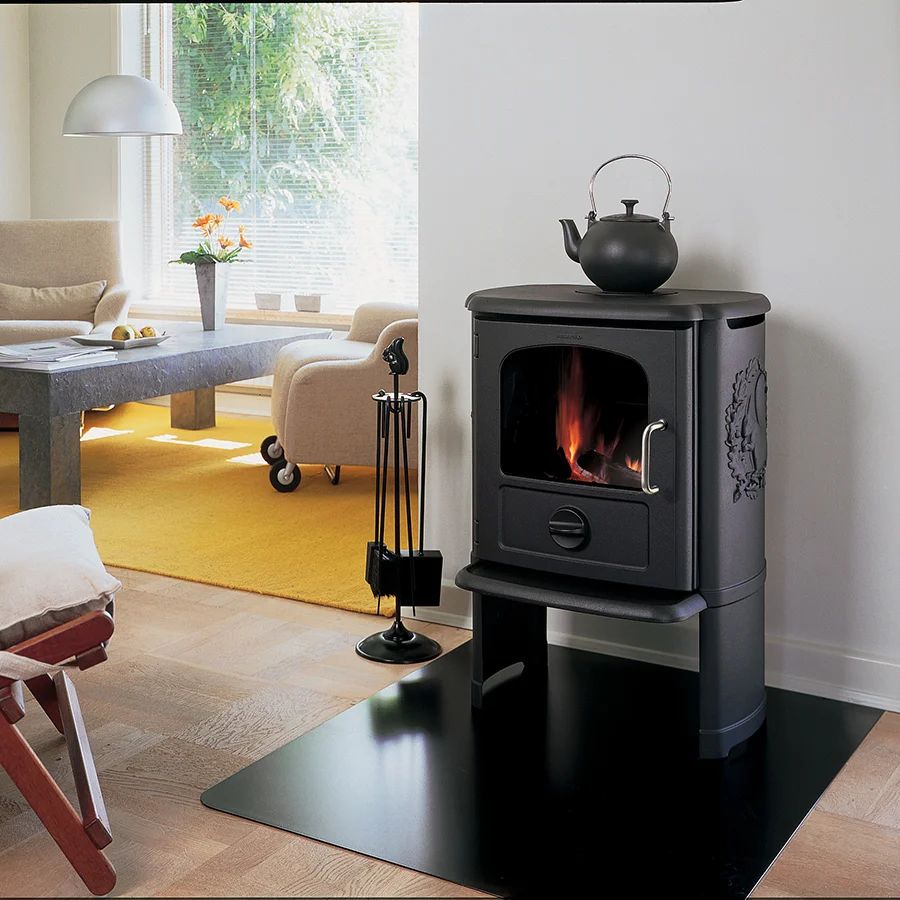 Classic black Morsø cast-iron wood stove with the iconic squirrel relief on its side and a kettle on top, lit in a warm room with a yellow rug