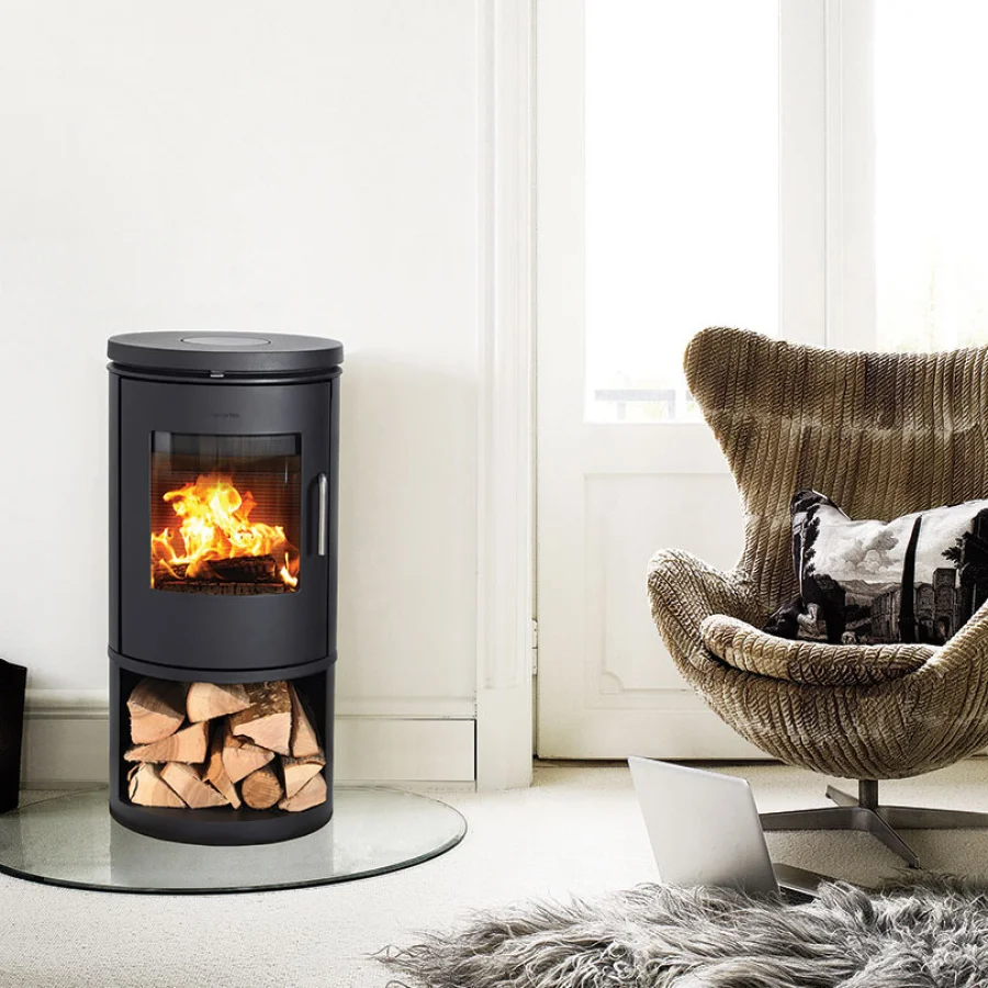 Contemporary black cylindrical Morsø wood stove with an integrated log store below, lit beside a modern egg chair in a bright room