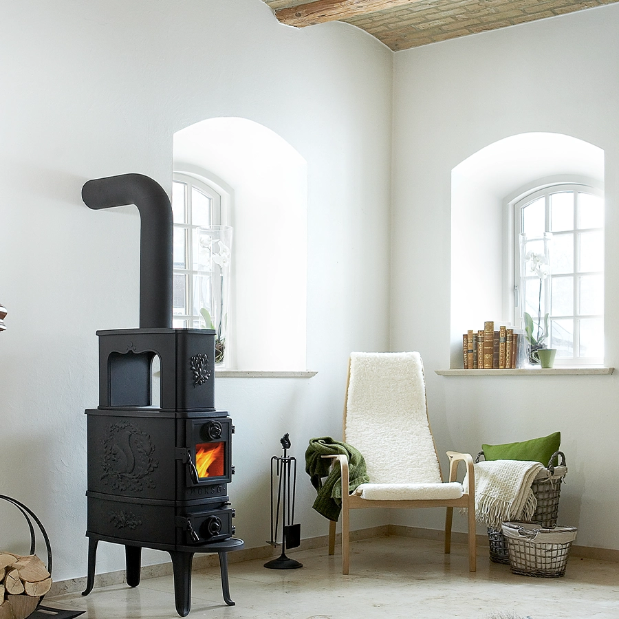 Tall classic Morsø cast-iron wood stove with a squirrel relief on its side, lit in a bright, whitewashed room with arched windows and a sheepskin chair