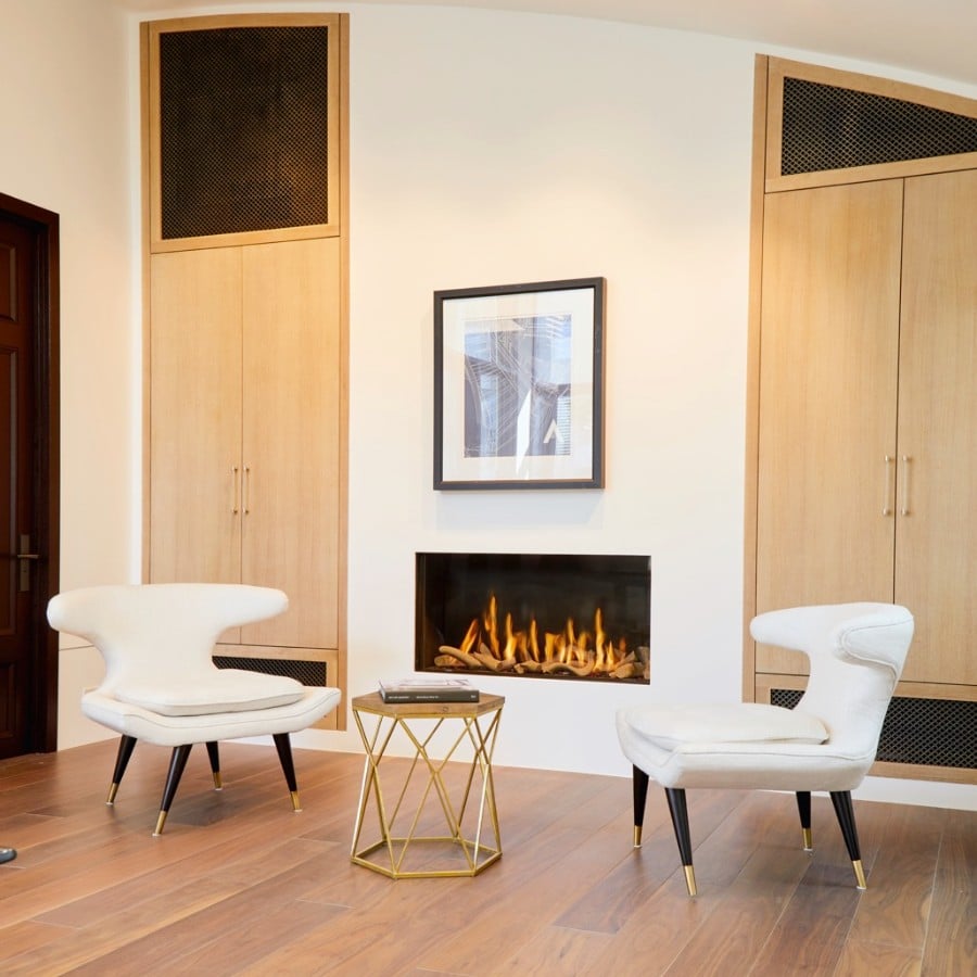 Ortal front-facing gas fireplace set into a white wall between tall oak cabinets with cane panels, a single framed photograph above the fire and a pair of sculptural white chairs in front