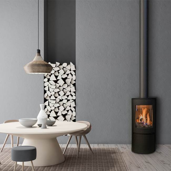 Element4 Ovation gas stove standing against a soft grey wall in a modern dining room, its flue rising the full height of the room, beside a pale round table with a woven pendant lamp and a recessed niche stacked with birch logs