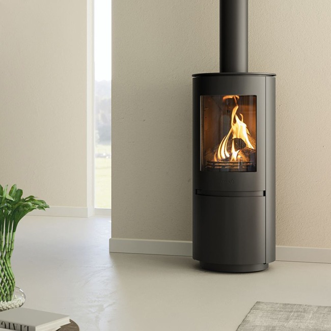 Element4 Ovation freestanding gas stove — a rounded black steel cylinder on a plinth with a tall portrait window of flame and a slim black flue rising to the ceiling — standing against a pale wall beside a floor-to-ceiling window looking out over green countryside