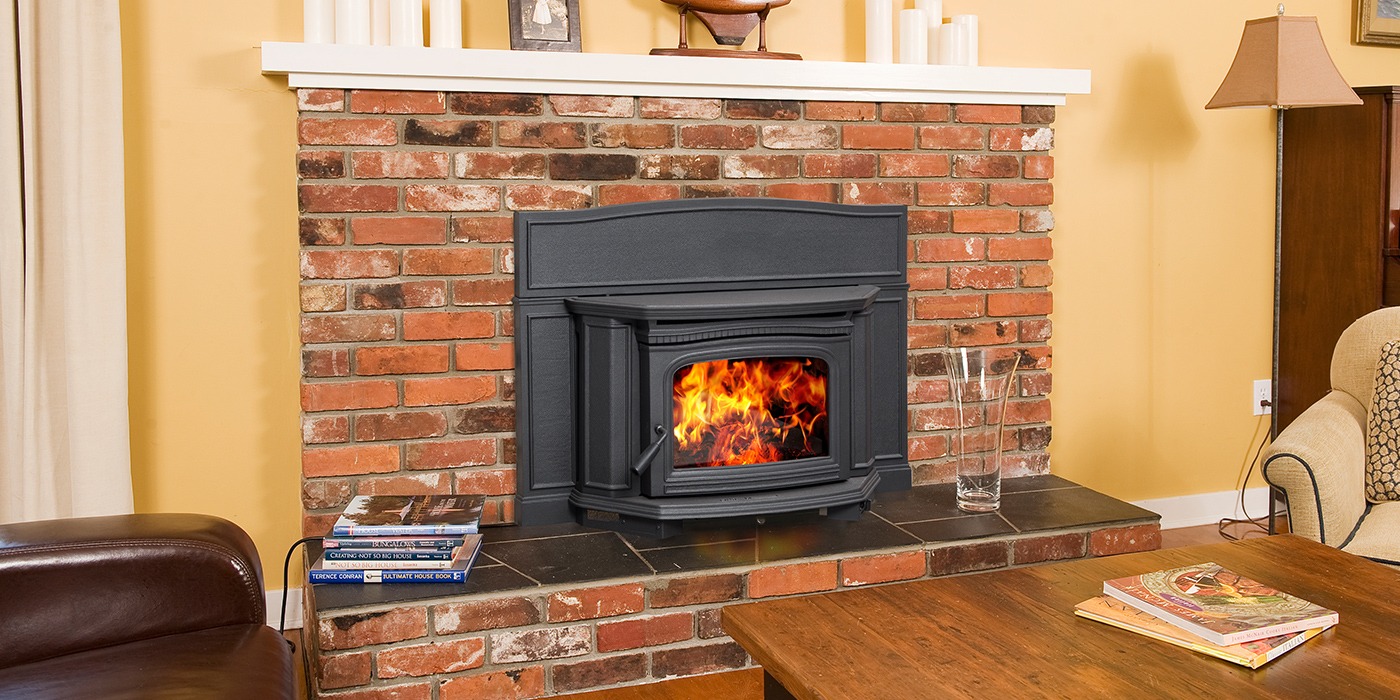 Pacific Energy Alderlea T5 Insert LE, a black cast-iron insert with an arched top and pilasters, set into a red brick fireplace beneath a white mantel in a yellow sitting room with a leather armchair and stacked books