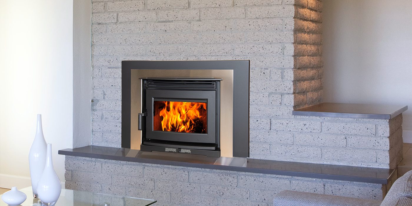 Pacific Energy Neo 1.6 Insert LE2 with a stainless-steel faceplate and black contemporary trim set into a grey brick fireplace, a polished steel hearth shelf running beneath it and white vases on a glass table