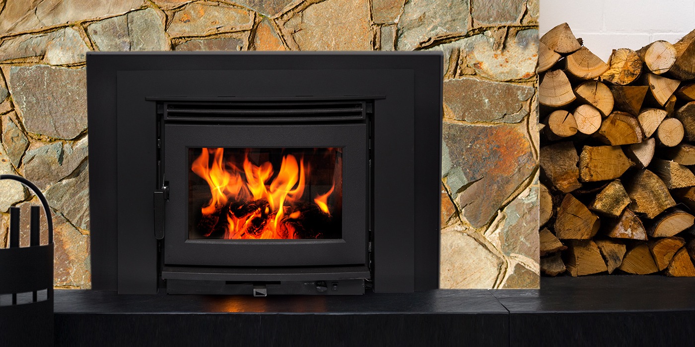 Pacific Energy Neo 2.5 Insert LE2 with a black faceplate set into a rust-toned slate fireplace above a black stone hearth, with split firewood stacked beside it