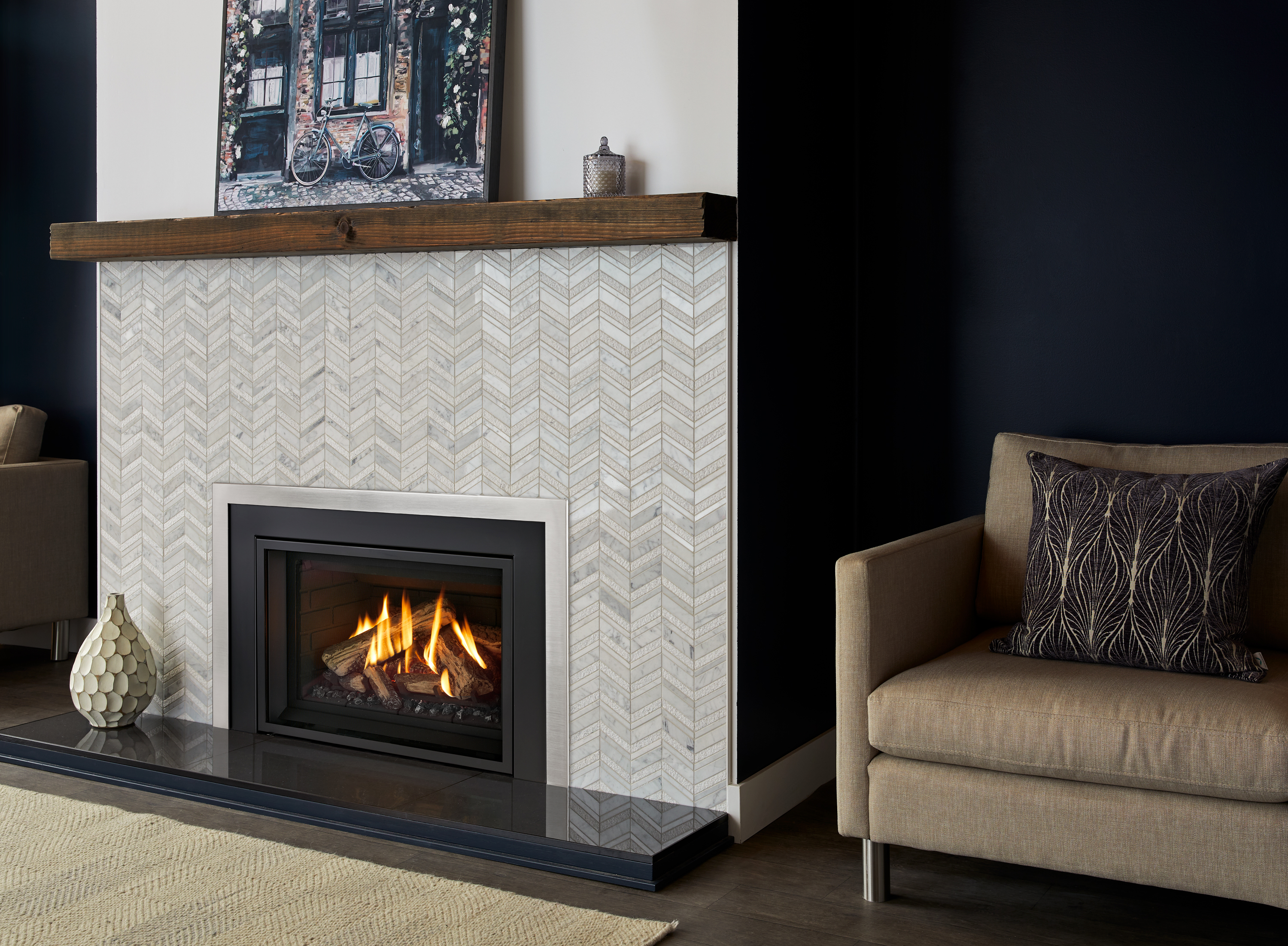 Regency Gi29LE gas insert with a glowing log fire, set into a grey chevron-tile reface with a reclaimed-wood mantel and black stone hearth, flanked by tan armchairs against a navy wall