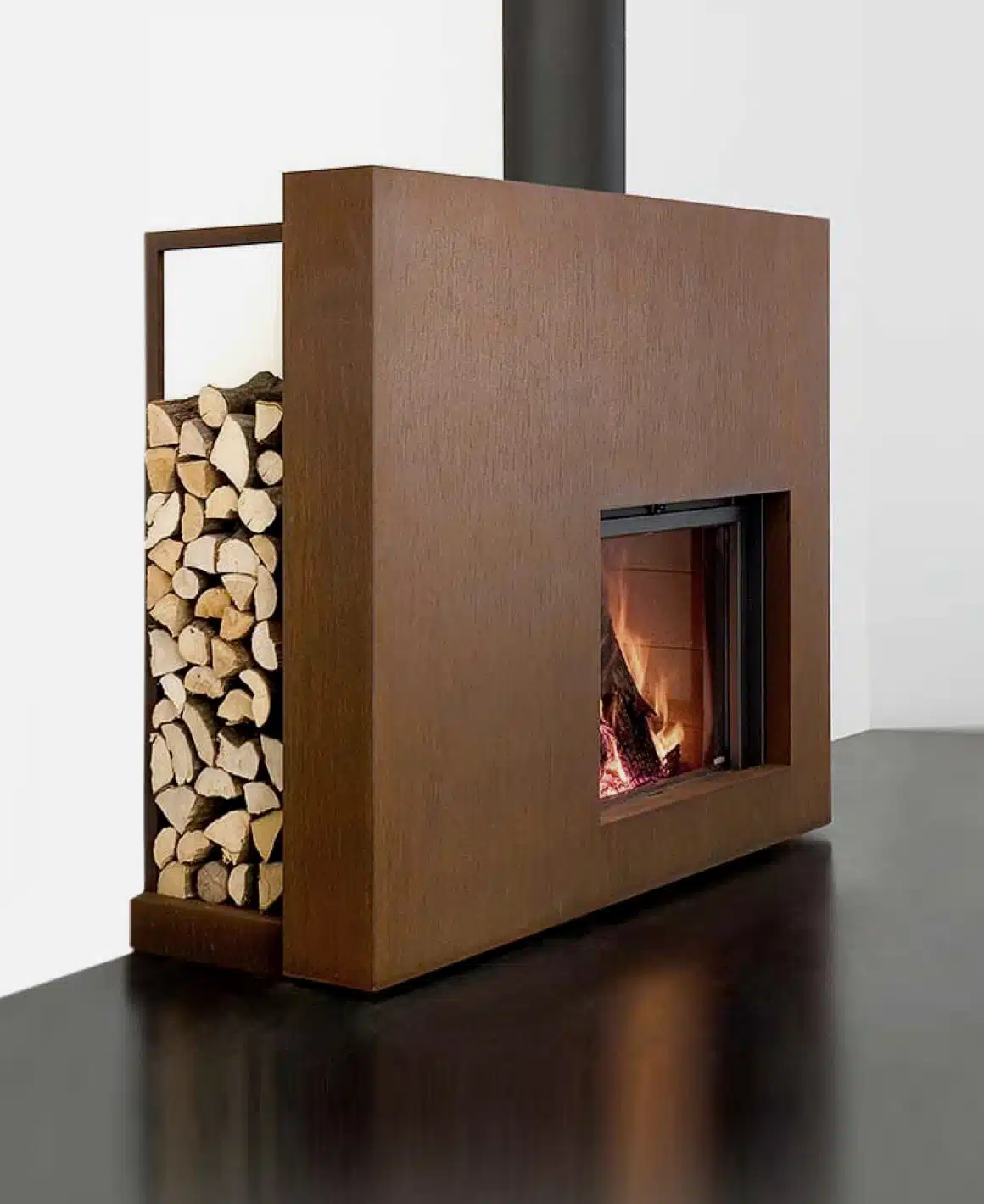 Stûv 21 Clad fireplace in a rust-brown weathering-steel finish with an integrated log store stacked with firewood alongside the fire, shown as a sculptural freestanding block against a white wall