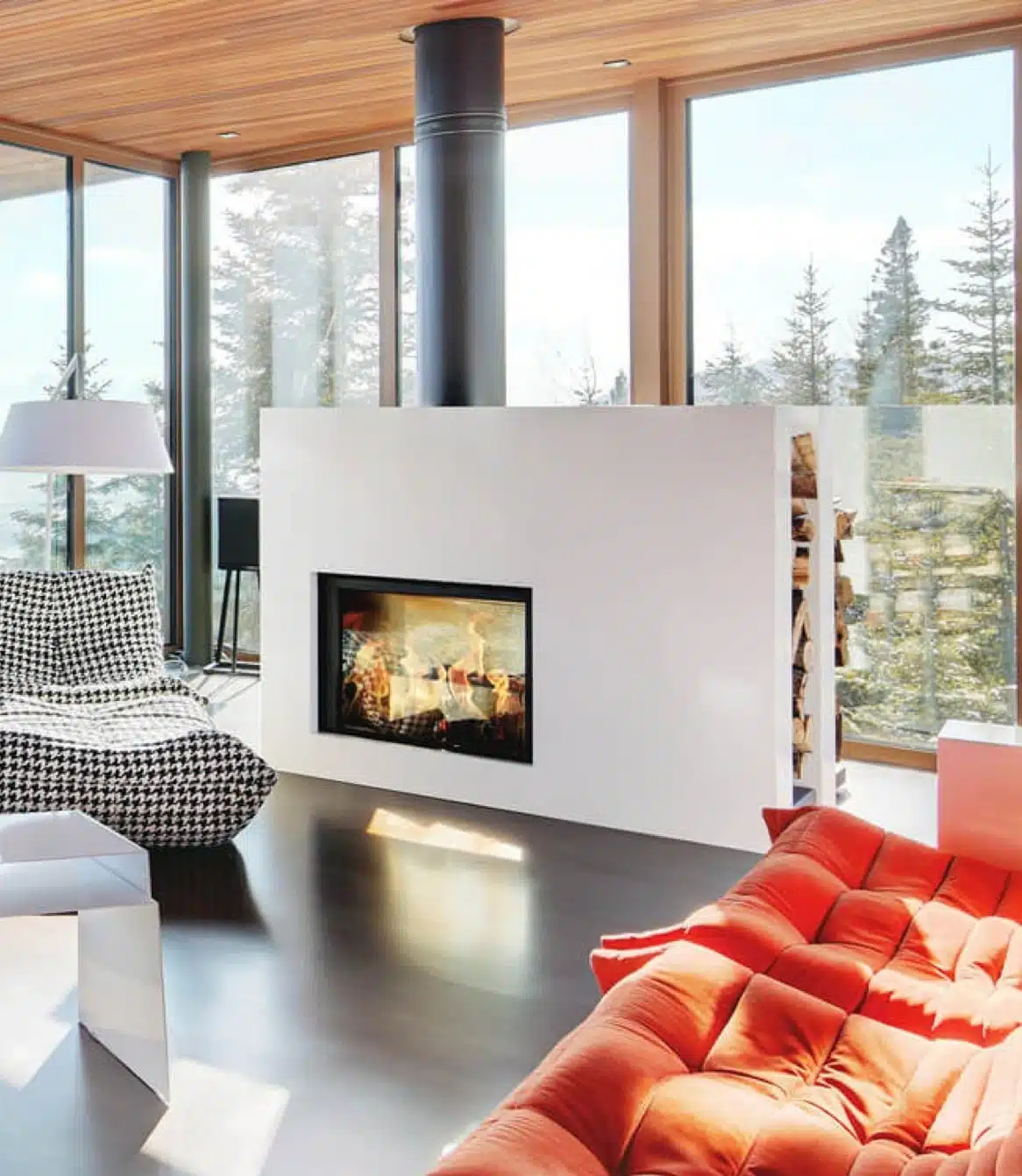Stûv 21 Clad freestanding wood fireplace in a white finish, standing as a room divider in a bright cabin with floor-to-ceiling forest views, a wood-plank ceiling and a red sofa