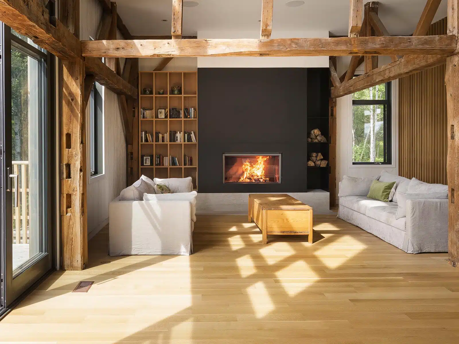 Stûv 21 single-faced wood fireplace set into a black feature wall in a converted timber-frame barn, flanked by a built-in bookshelf and a firewood niche, with reclaimed beams overhead and sunlight across a pale wood floor
