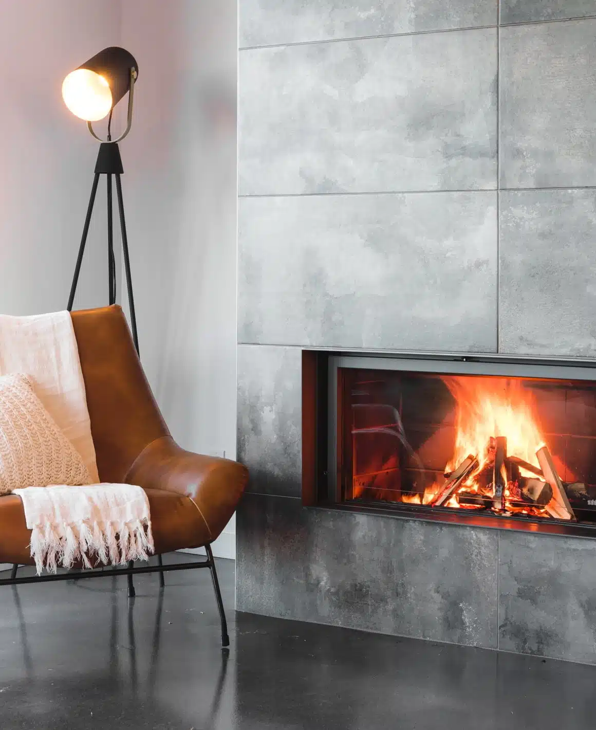 Stûv 21 single-faced wood fireplace set into a grey concrete-tile wall, a wide linear fire behind frameless glass, beside a tan leather chair and a tripod lamp in a calm modern room