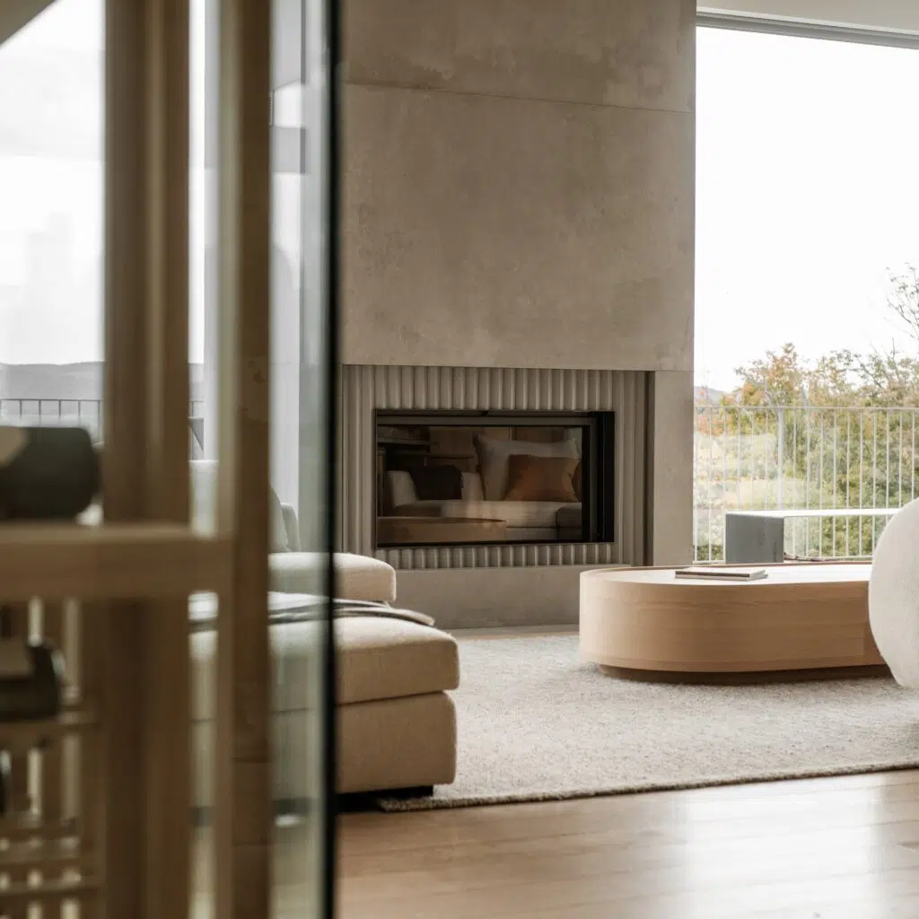 Stûv 21 wood fireplace in a fluted concrete surround, seen through a doorway in a bright minimalist living room with a round wood coffee table and a view of autumn trees