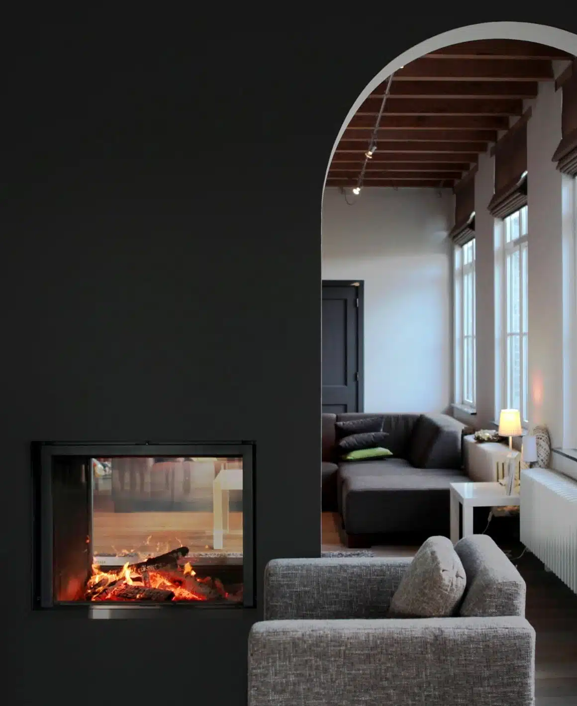 Stûv 21 double-faced see-through wood fireplace built into a charcoal wall, the fire visible from a sitting room and from the lounge beyond through an arched opening