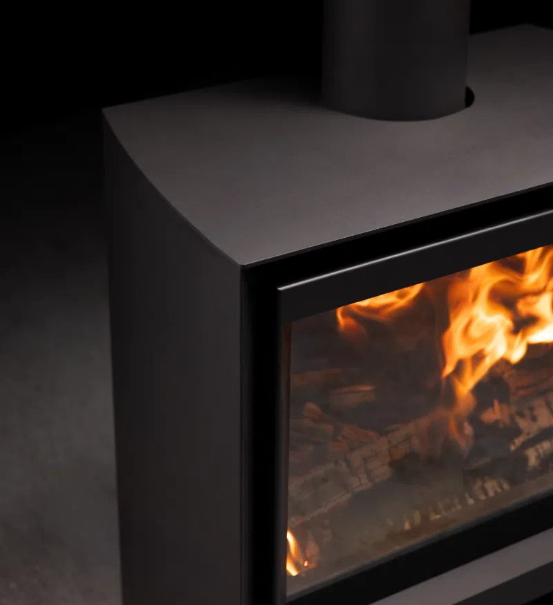Close view of the Stûv 6-H&rsquo;s upper corner — the black steel body visibly narrower at the back and widening toward the door frame, its top edge softened into a curve where the flue leaves it, with flames filling the glass below