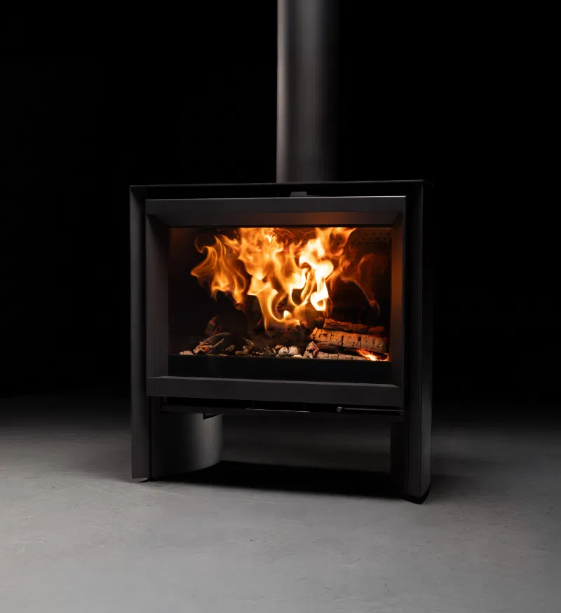 The Stûv 6-H seen head-on in a darkened studio — a compact black wood stove raised on its own slim legs, an open recess beneath the firebox, and tall flames filling the full width of the glass door