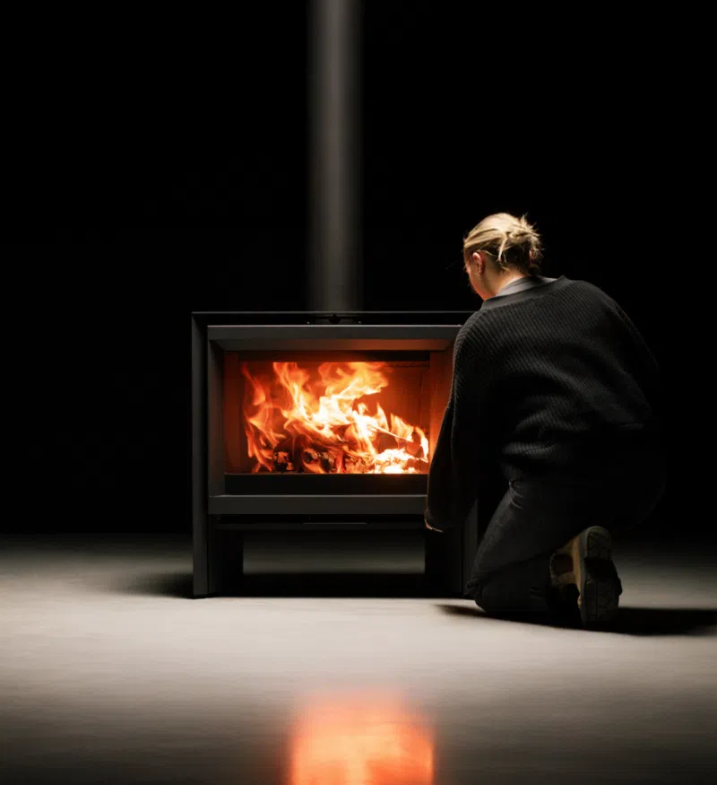 A woman kneeling beside a lit Stûv 6-H in a darkened room, tending the fire — the stove&rsquo;s wide glass door full of flame, its light thrown across the floor, and a slim black flue rising into the dark above