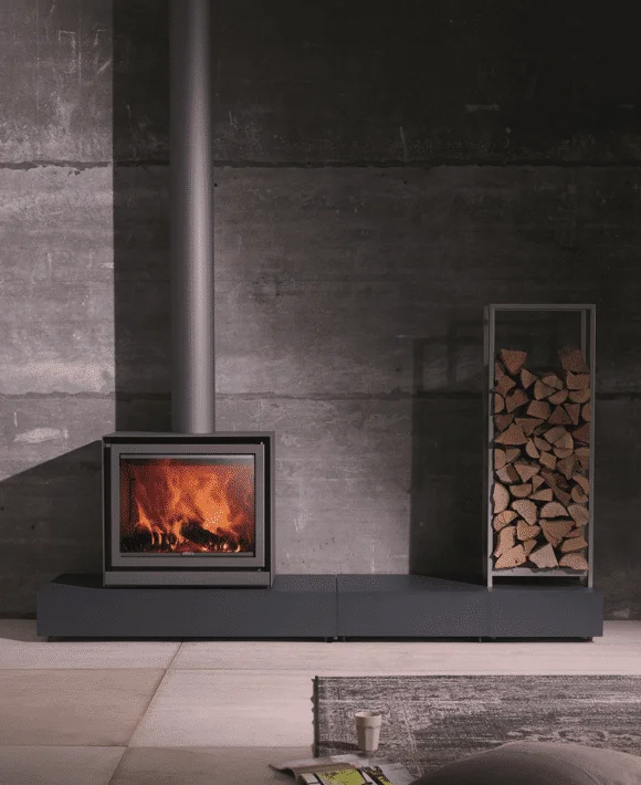 Stûv 16 Cube wood stove on a long podium against a concrete wall with a vertical log rack alongside