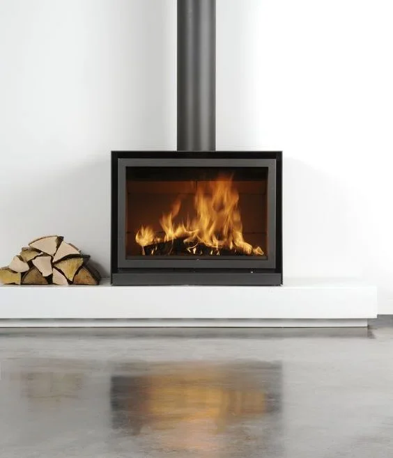 Stûv 16 Cube wood stove with a roaring fire on a clean white plinth, birch logs stacked alongside, polished concrete floor reflecting the flames