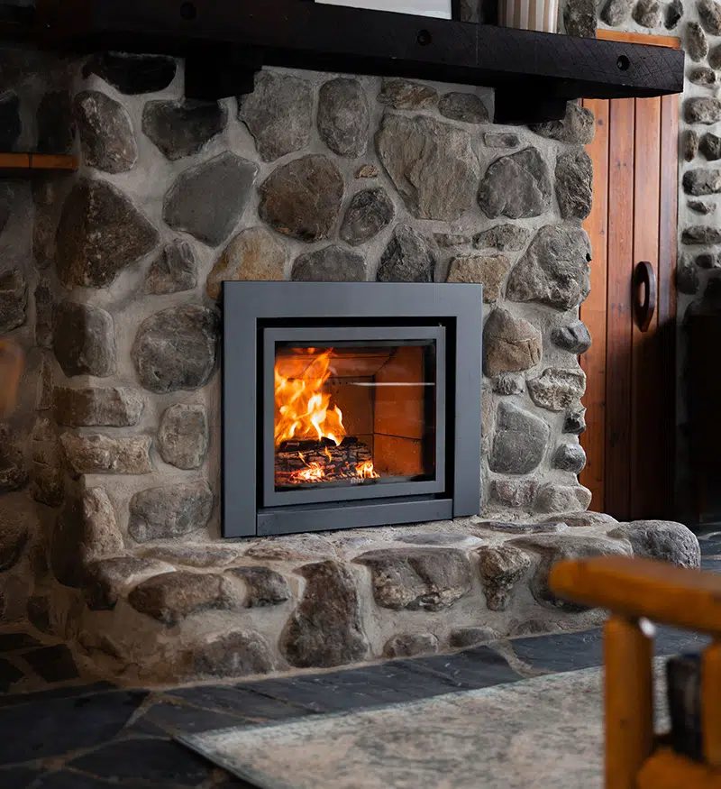 Stûv 16-In wood-burning insert retrofit into a rustic stone masonry fireplace with a dark wood mantle beam and slate hearth, fire burning inside