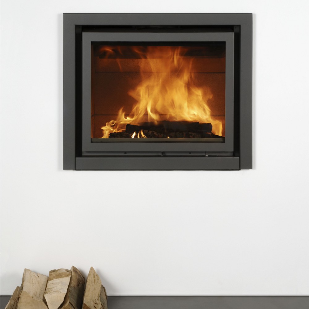Stûv 16-In wood-burning insert set flush into a clean white wall with a black steel surround, a strong fire behind the glass and split firewood stacked below